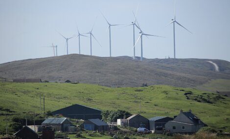 A series of wind turbines on a grassy hill with several houses and buildings in the foreground.