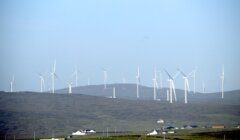 Several wind turbines spread across a hilly landscape under a clear sky, with a few buildings visible in the foreground.