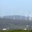 Several wind turbines spread across a hilly landscape under a clear sky, with a few buildings visible in the foreground.