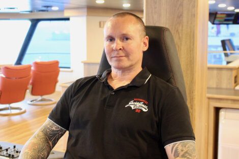 A man with tattoos sits in a chair on a ship's bridge wearing a black shirt with a red and white logo. The background shows orange chairs and large windows.