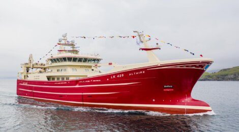 A large red and white fishing vessel named "LK 429 ALTAIRE" is decorated with colorful flags and positioned on calm waters against an overcast sky.