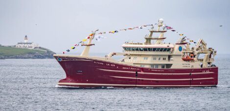 A large red and white fishing vessel named "ALTAIRE LK 429" adorned with flags sails on the sea, with a lighthouse on the coastline in the background.