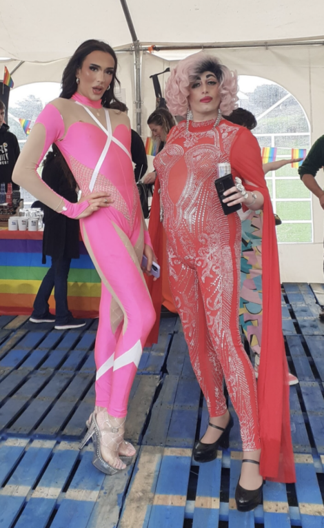 Two people in colorful drag outfits pose under a tent; one in a pink jumpsuit with high heels, the other in a red jumpsuit with a red cape, holding a drink. Rainbow flags are in the background.