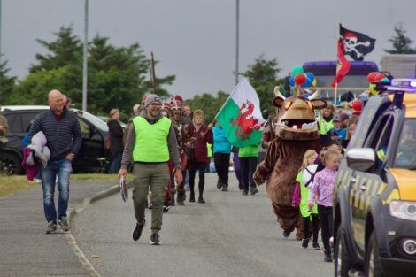 A group of people participate in a festive parade, featuring a person in a furry costume, colorful balloons, a dragon flag, and onlookers. A police vehicle is nearby.