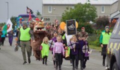 A parade featuring children in costumes, holding shields and signs, led by adults in neon vests. A person in a large costume resembling a fictional creature is also part of the parade.