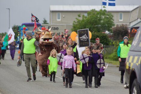 A parade featuring children in costumes, holding shields and signs, led by adults in neon vests. A person in a large costume resembling a fictional creature is also part of the parade.