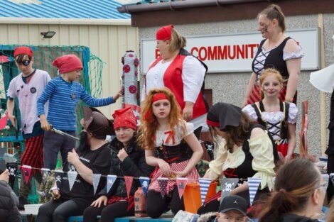 A group of children and adults in pirate costumes participate in a community event, standing and sitting on a float decorated with pirate-themed banners.