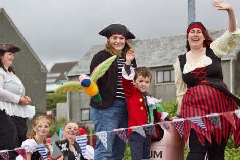 A group of people dressed in pirate costumes, including children and adults, smile and wave while standing on a decorated float with bunting in a neighborhood.