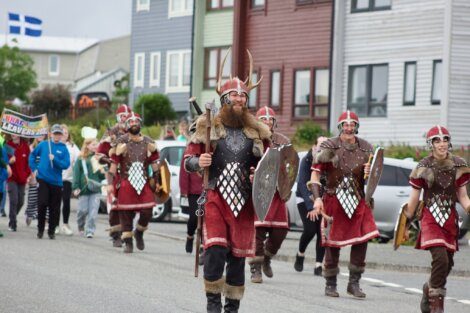 People dressed in Viking costumes, including helmets and shields, march down a street. Colorful houses are in the background, and other people follow the costume group.