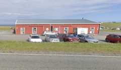 A red building with a grey roof sits behind a row of parked cars on a clear day. The building has multiple windows and doors, and the surrounding area features grass and a road in the foreground.