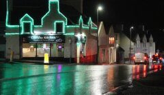 A street at night with lit buildings, including a restaurant with neon green lights named "Gurkha Kitchen." The wet road reflects the lights, and vehicles are visible in the background.