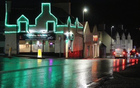 A street at night with lit buildings, including a restaurant with neon green lights named "Gurkha Kitchen." The wet road reflects the lights, and vehicles are visible in the background.