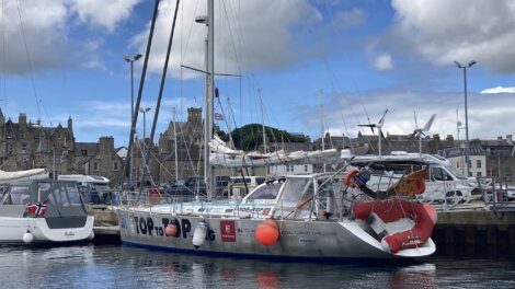 A sailboat named "TOPtoTOP" docked in a harbor, equipped with a red lifebuoy and wrapped with promotional graphics. Other boats and historic stone buildings are visible in the background.