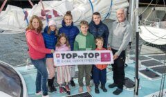 A group of eight people, including children and adults, stand together on a boat holding a banner that reads "TOPtoTOP Global Climate Expedition." They are surrounded by ropes and sails.