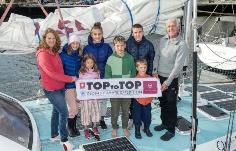 A group of eight people, including children and adults, stand together on a boat holding a banner that reads "TOPtoTOP Global Climate Expedition." They are surrounded by ropes and sails.