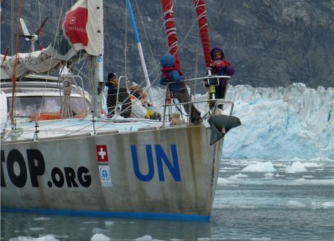 A sailboat with "UN" and "OOP.org" logos navigates icy waters near a glacier, with several crew members on board dressed in cold-weather gear.