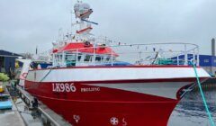 A red and white fishing vessel docked beside a pier with the identification "LX986 Prolific." Various equipment is visible on the boat, and colorful flags are strung along the mast.