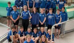 A group of young swimmers wearing blue uniforms poses for a team photo at an indoor pool facility. Some are standing while others sit on the edge of the pool area.