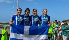 Four athletes in blue and white uniforms hold a blue and white flag, displaying medals around their necks. They stand outdoors with spectators in the background.