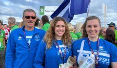 Three people wearing blue uniforms stand together at an outdoor event, holding a white flag and an award. Other participants and a banner are visible in the background.