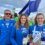 Three people wearing blue uniforms stand together at an outdoor event, holding a white flag and an award. Other participants and a banner are visible in the background.