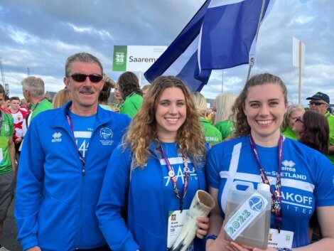 Three people wearing blue uniforms stand together at an outdoor event, holding a white flag and an award. Other participants and a banner are visible in the background.