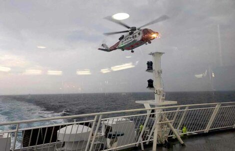 A red and white helicopter hovers above a ship's deck at sea with cloudy skies in the background.