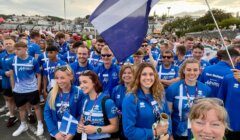 A group of people in blue uniforms stand together, some holding flags. They are outdoors in a crowd, and a woman in a green shirt is in the foreground taking a selfie.