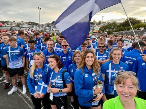 A group of people in blue uniforms stand together, some holding flags. They are outdoors in a crowd, and a woman in a green shirt is in the foreground taking a selfie.