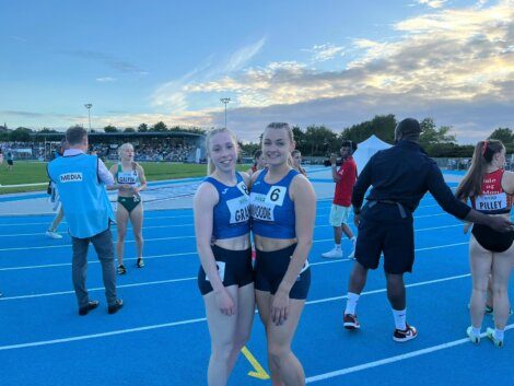 Two female athletes wearing blue athletic uniforms stand on a blue track, smiling at the camera. Other people and athletes are in the background, along with a cloudy sky at sunset.