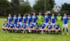 A group of soccer players in blue and white uniforms poses for a team photo on a grassy field, with trees and houses in the background.