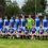 A group of soccer players in blue and white uniforms poses for a team photo on a grassy field, with trees and houses in the background.