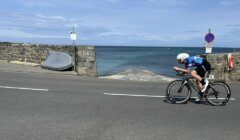 A cyclist in blue attire rides a road bike near a stone wall with water in the background under a partly cloudy sky. A sign is visible next to the cyclist and a boat is positioned by the wall.