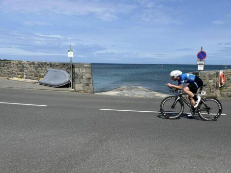 A cyclist in blue attire rides a road bike near a stone wall with water in the background under a partly cloudy sky. A sign is visible next to the cyclist and a boat is positioned by the wall.