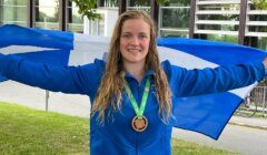 A person with wet hair, wearing a blue jacket, holds a blue and white flag while smiling. They have a medal around their neck and are standing outdoors with a building in the background.