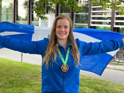 A person with wet hair, wearing a blue jacket, holds a blue and white flag while smiling. They have a medal around their neck and are standing outdoors with a building in the background.