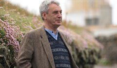 A man in a tweed jacket stands with hands in pockets beside a stone wall with blooming flowers, with a blurred lighthouse in the background.