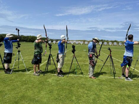 A group of people standing on a grassy field, aiming recurve bows at targets set up in the distance under a clear blue sky. Tripods are placed beside some archers.