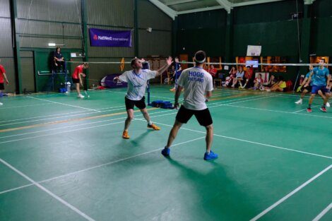 Indoor badminton game in progress. A player in a white shirt prepares to hit the shuttlecock while another player in a blue shirt and black shorts stands ready. Spectators watch from the sideline.