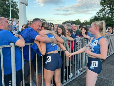 Athletes wearing competition numbers 6 and 8 stand next to a metal barrier; number 6 hugs a man, while number 8 holds hands with number 6. Spectators watch from the other side of the barrier.