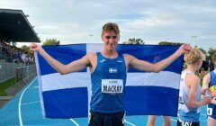 Athlete on a track holds up a flag behind him. His race bib reads "MACKAY." Other athletes and spectators are visible in the background.