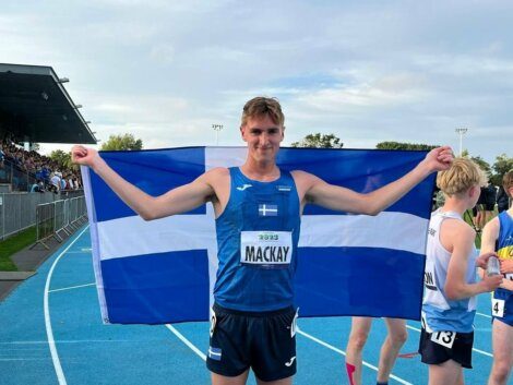 Athlete on a track holds up a flag behind him. His race bib reads "MACKAY." Other athletes and spectators are visible in the background.