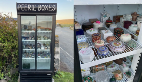 A glass-front refrigerator labeled "Peerie Boxes" displaying various decorative, colorful small boxes on shelves inside. An outdoor scene with greenery and a road is visible in the background.