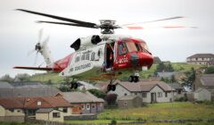 A red and white Coastguard rescue helicopter is in flight above a residential area with houses in the background.