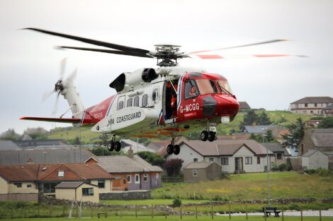 A red and white Coastguard rescue helicopter is in flight above a residential area with houses in the background.