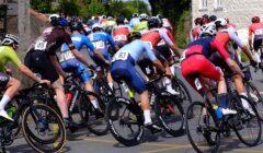 Cyclists in colorful jerseys are riding closely together on a road during a race. The background includes greenery and buildings.