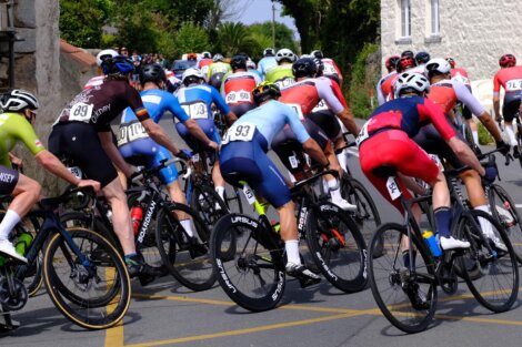 Cyclists in colorful jerseys are riding closely together on a road during a race. The background includes greenery and buildings.