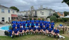 A group of 20 men in blue sports jerseys pose for a team photo outside Les Douvres Hotel. The background features hotel buildings, a swimming pool, and a grassy area with trees.