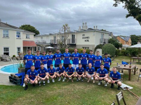 A group of 20 men in blue sports jerseys pose for a team photo outside Les Douvres Hotel. The background features hotel buildings, a swimming pool, and a grassy area with trees.