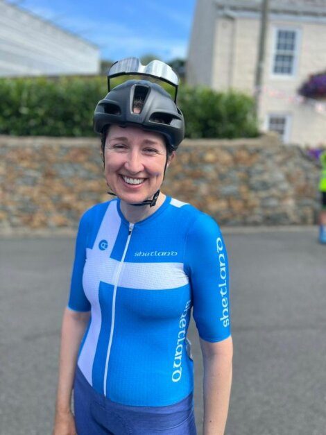 A cyclist in a blue and white kit stands smiling, wearing a helmet with sunglasses on top. A stone wall and buildings are in the background.